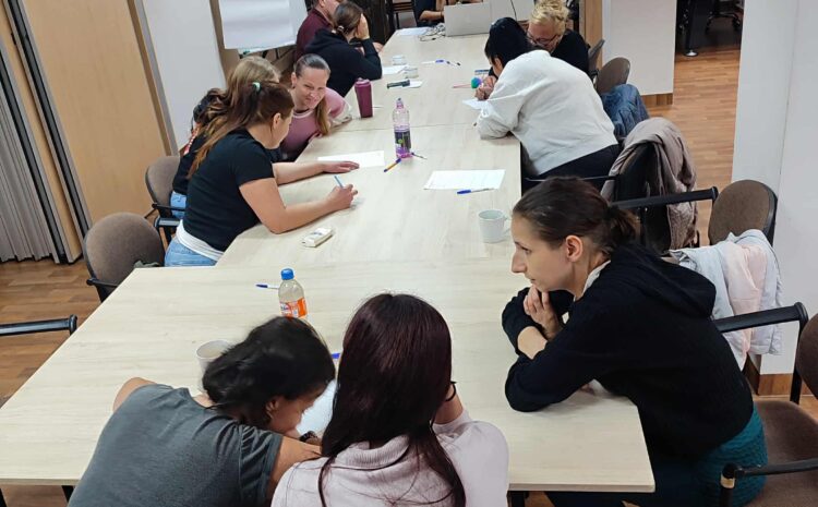 Na zdjęciu widać grupę uczestników siedzących przy dużym stole podczas warsztatów. W tle prowadząca omawia materiał wyświetlony na ekranie.