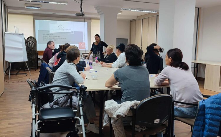 Na zdjęciu widać grupę uczestników siedzących przy stole podczas warsztatów. Z boku widoczny jest chodzik, a cała scena pokazuje spokojną, roboczą atmosferę spotkania.