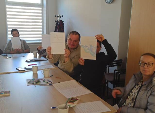 Na zdjęciu uczestnicy warsztatów siedzą przy stole i prezentują swoje prace - rysunki fal, które symbolizują dzieci, emocje i samych siebie. Atmosfera jest swobodna i twórcza.