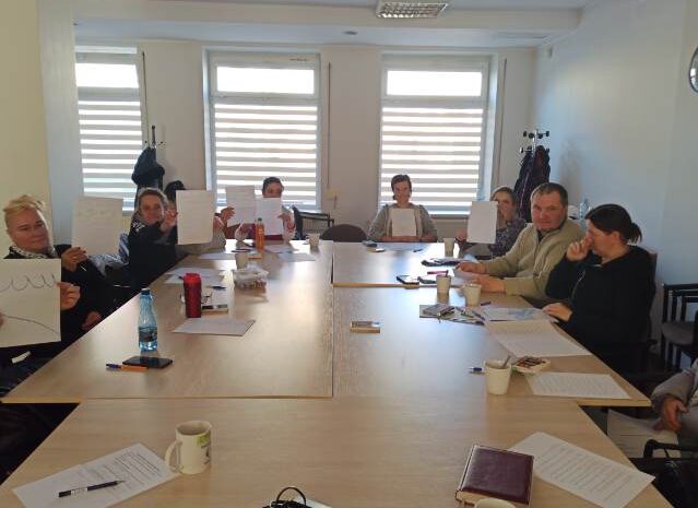 Na zdjęciu uczestnicy warsztatów siedzą wokół stołu i trzymają w dłoniach swoje rysunki fal - wykonane w ramach ćwiczenia symbolicznego, odnoszącego się do dzieci, emocji i własnych przeżyć.