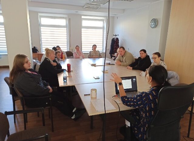 Na zdjęciu widać uczestników warsztatów siedzących wokół dużego stołu i słuchających prowadzącej. Grupa jest skupiona, atmosfera wygląda na spokojną i sprzyjająca rozmowie.