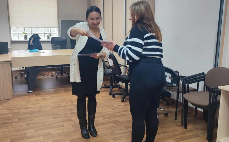 Na zdjęciu prowadząca przekazuje uczestniczce dyplom. Obie stoją w sali warsztatowej uśmiechnięte i zaangażowane. Scena przedstawia zakończenie udziału w zajęciach.